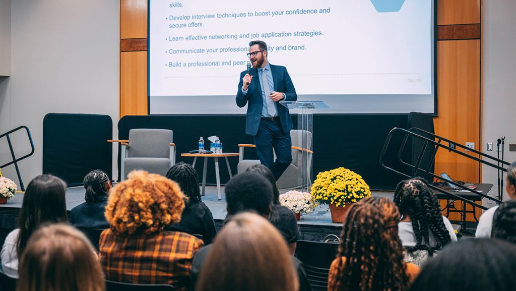 Speaker crosses stage in front of a screen with career prep tips as students look on from the audience.