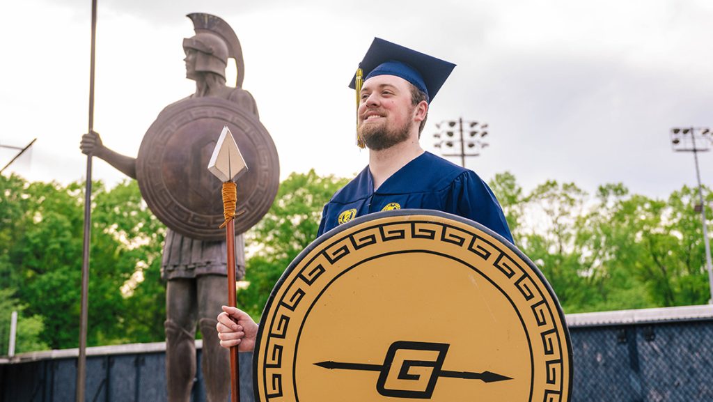 Graduate with Spiro shield and arrow stands in front of the Spiro statue.