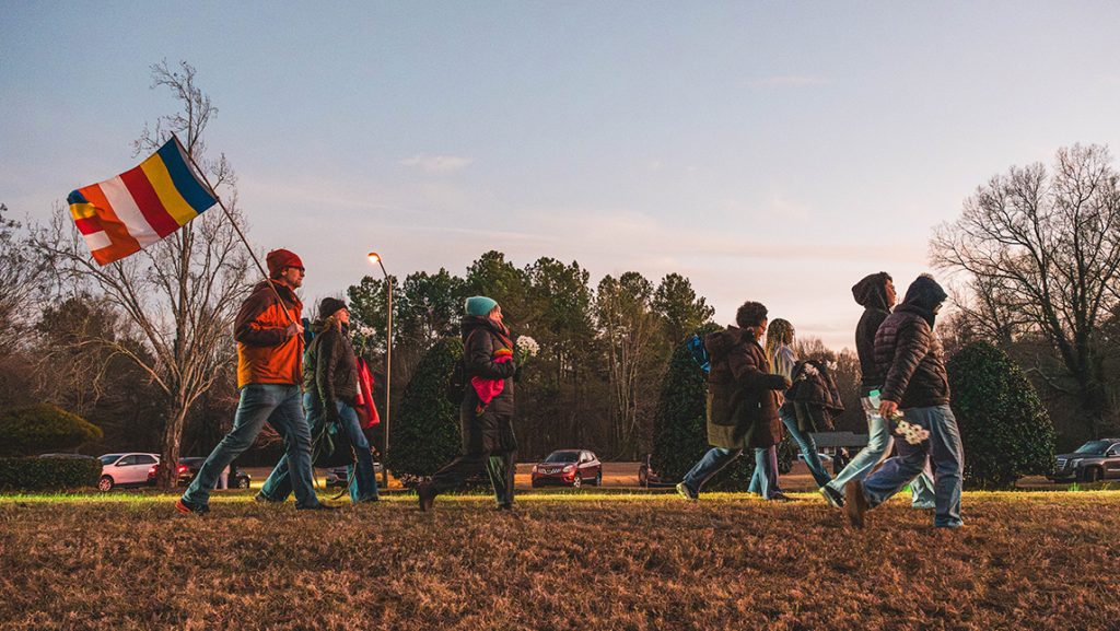 Students walk at dusk with traffic behind them. The faculty member at the back carries a Buddhist flag.