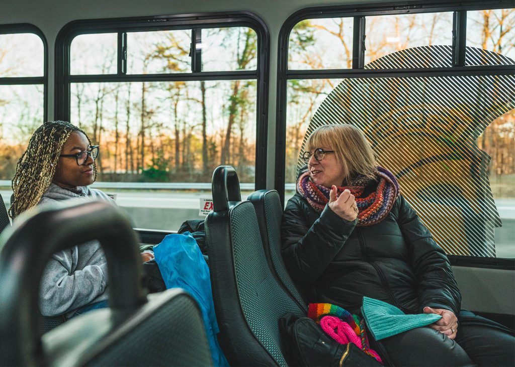 Student and teacher talk as they ride on a bus with the Spartan head decal on the window behind them.