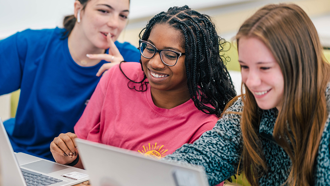 UNCG students look over one another's shoulders at a laptop screen.