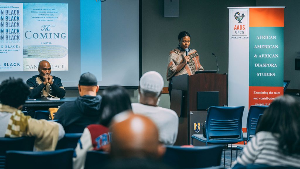 Speaker stands at a podium in front of an audience with an AADS banner in the background. Looks like they're discussing a book and the author is seated in traditional African clothes.