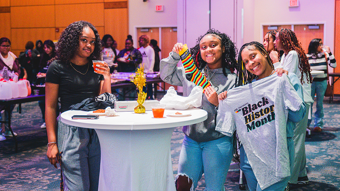 Three students stand around a high top table in a ballroom with refreshments and BHM t-shirts.