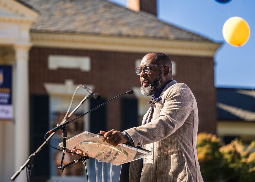 Man speaks at a podium outside of the Alumni house.