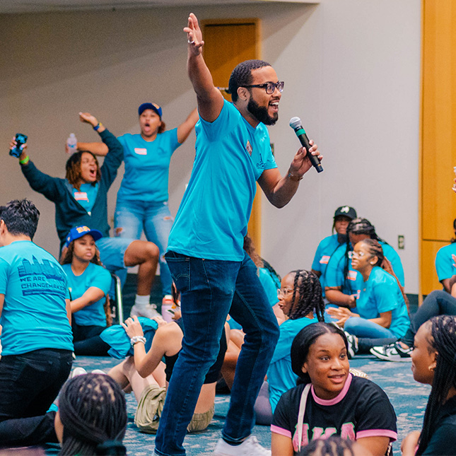 Man with a mic jumps excitedly surrounded by cheering students in the same blue t-shirt.
