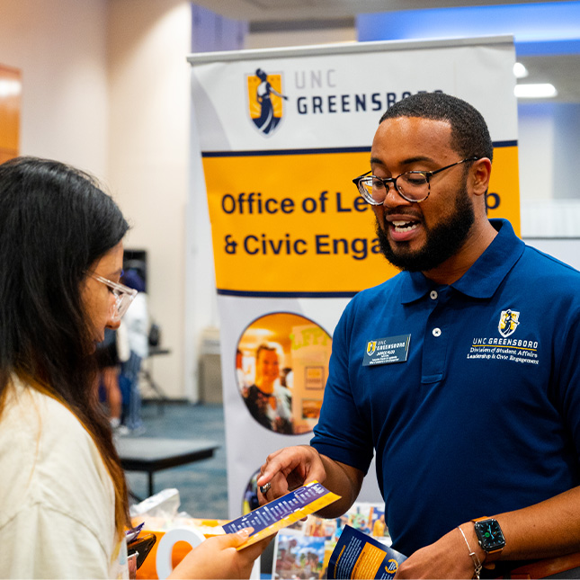 UNCG staff member speaks to a student in front of an OLCE banner. 