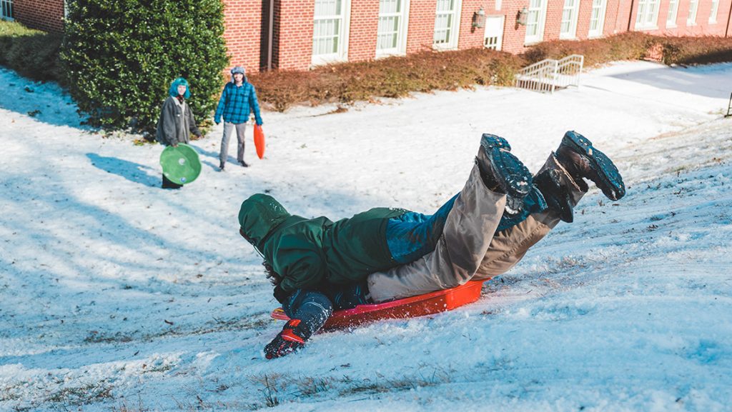 Student sleds down hill on campus on his belly while others watch at the bottom of the hill.