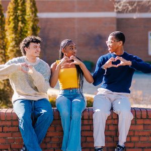UNCG students form hearts with their hands while sitting outside.