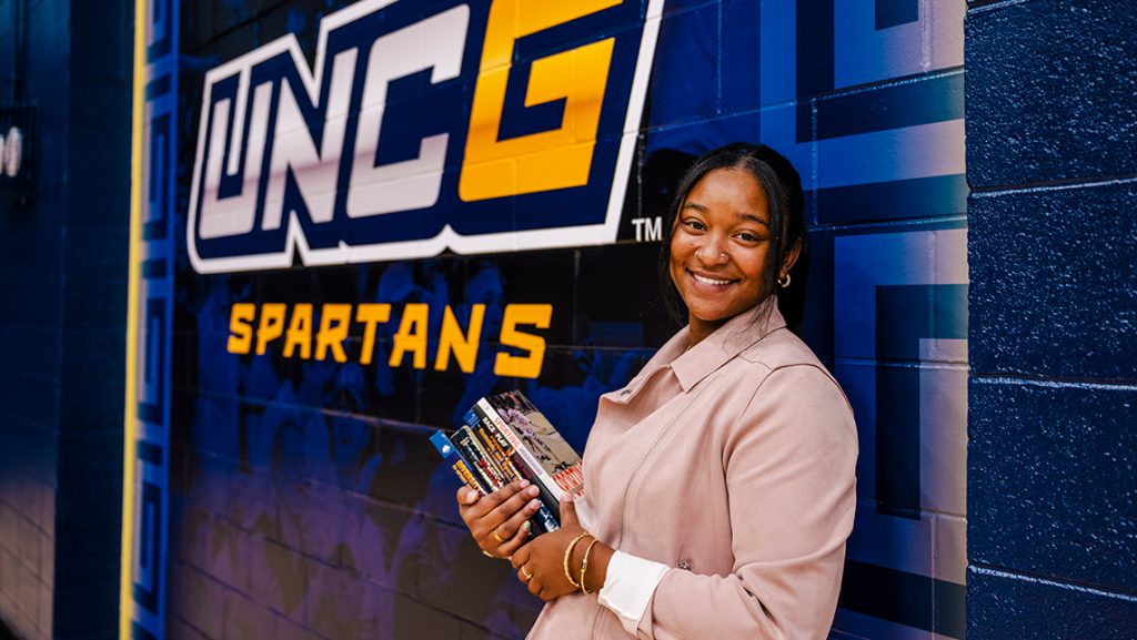 Student with books leans on a wall painted with UNCG Spartans.
