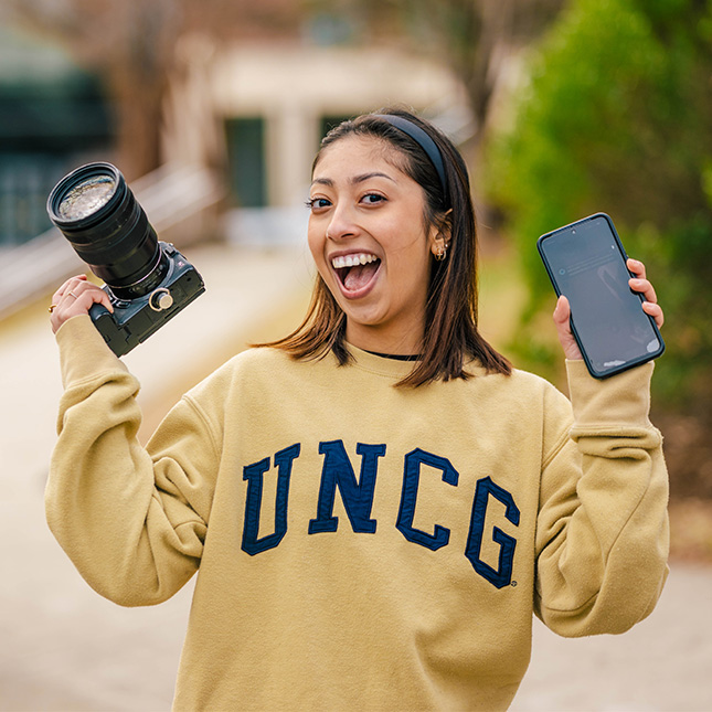 Student stands on campus and holds a DSLR camera in one hand and a cell phone in the other. 