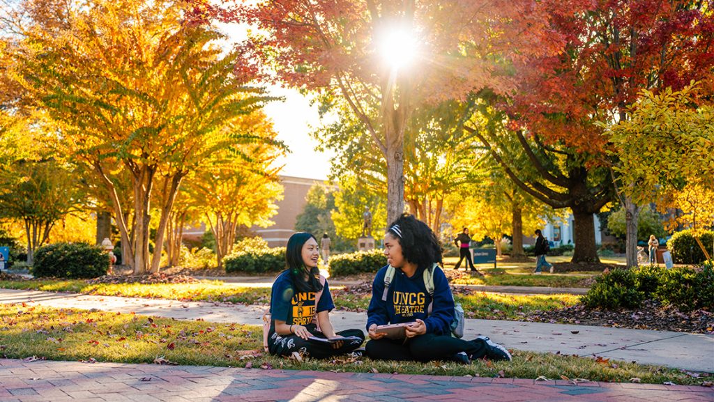 Two students sit in the quad on campus with brick classroom buildings and sunlight streaming through the fall trees behind them.