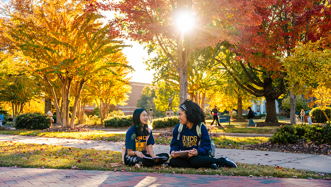 Two students sit in the quad on campus with brick classroom buildings and sunlight streaming through the fall trees behind them.