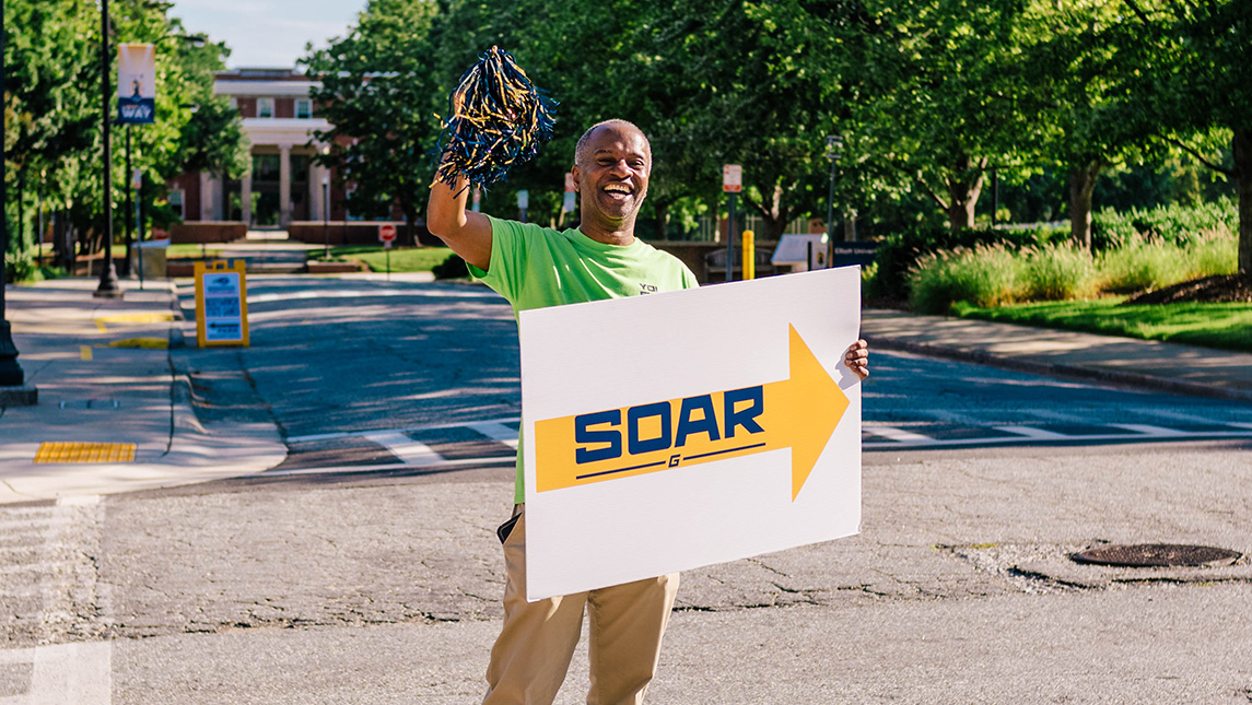 A UNCG employee holds a SOAR sign and waves a pom-pom.