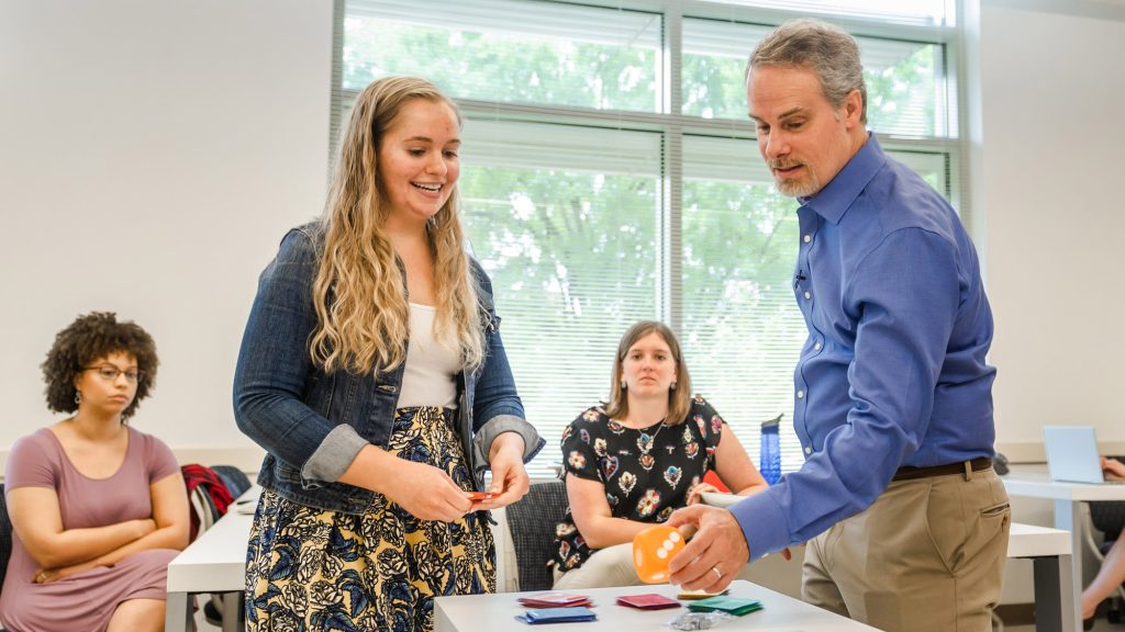A student and instructor interact over a game or activity on a table in a bright classroom while other students look on.