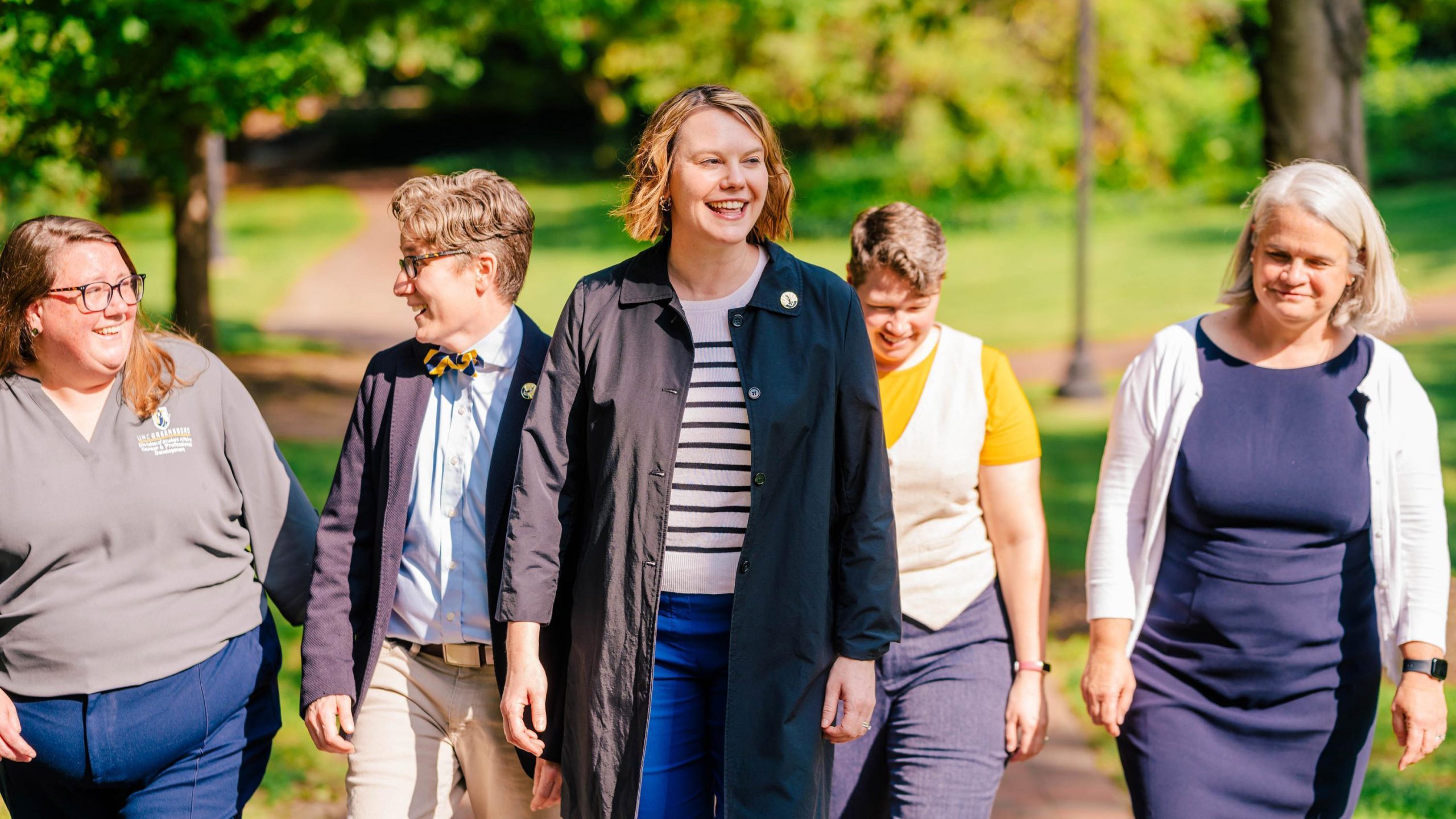 Five people walk together along a sunlit outdoor path surrounded by lush green trees and grass. They are smiling and laughing, conveying a sense of camaraderie.