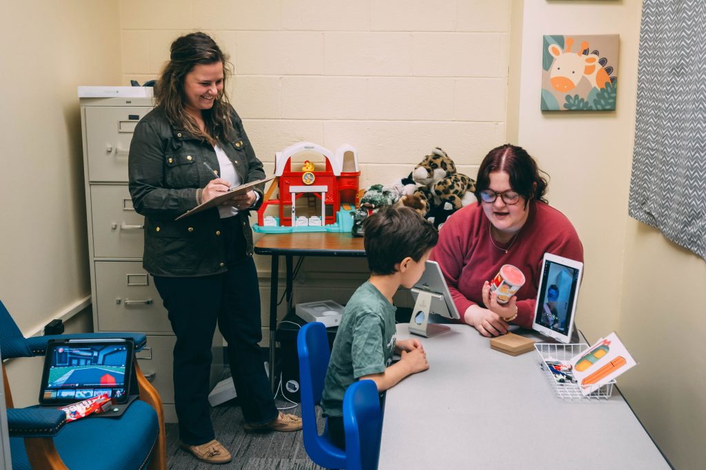 An undergraduate researcher interacts with a child, while a faculty member looks on. 