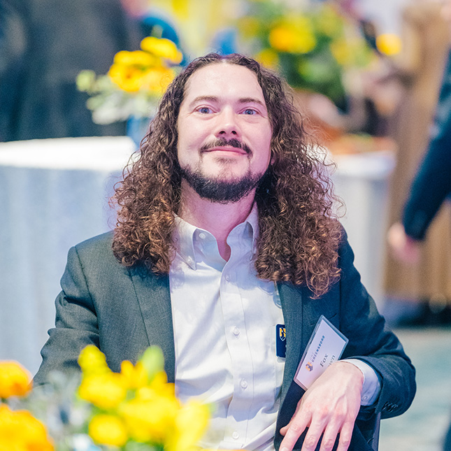 Student in a sports coat sits at a table with yellow centerpieces and smiles at the camera.