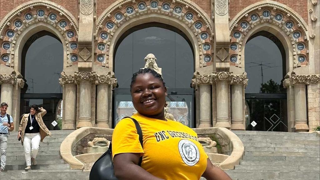 a woman in a UNCG yellow t-shirt stands in front of a statue