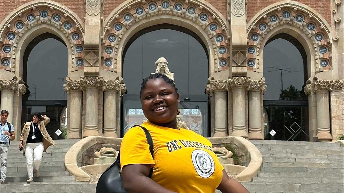 a woman in a UNCG yellow t-shirt stands in front of a statue
