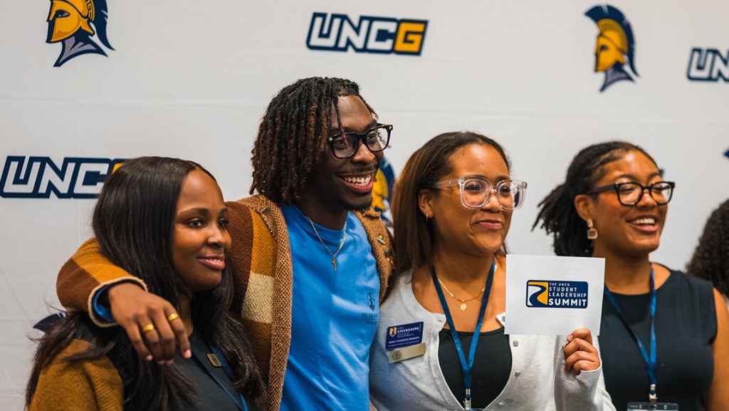 Four students stand in front of a screen with UNCG logo holding a Student Leadership Summit card.