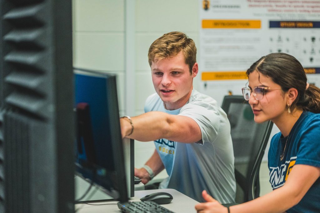 Two students gaze at a monitor in a lab, with one student pointing to something on the screen.