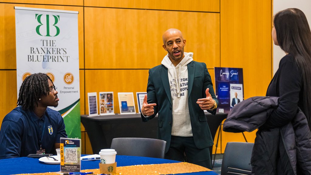 Professional speaks to two students with a Banker's Blueprint banner behind them.