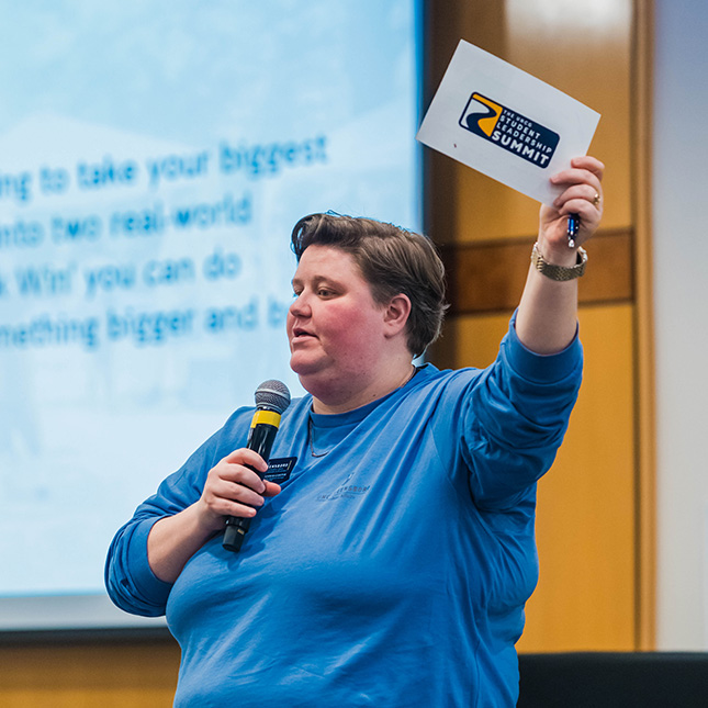 Staff member addresses a crowd with a mic and holds up a Student Leadership Summit card.