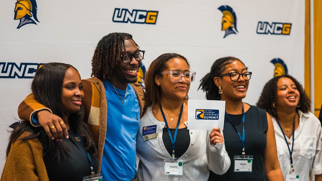 Students stand in front of a screen with UNCG logo holding a Student Leadership Summit card.