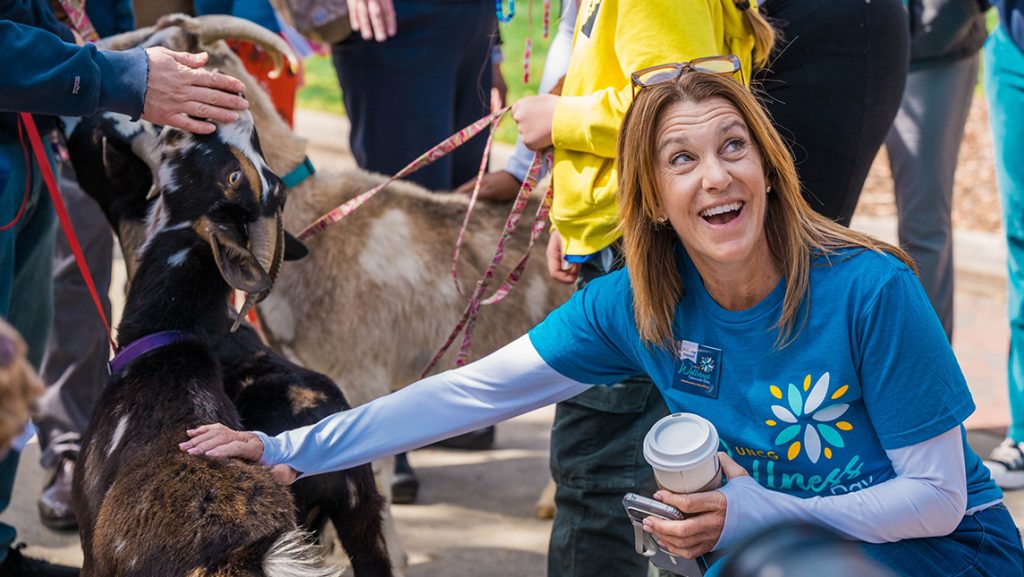 Staff member in wellness takeover t-shirt kneels to pet a goat and smiles.
