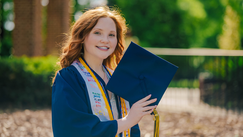 AnnaGrace Berry with red hair stands outdoors in a cap and gown, holding her mortarboard. She wears honor cords and a decorated stole, with trees and a sunlit campus walkway blurred in the background.