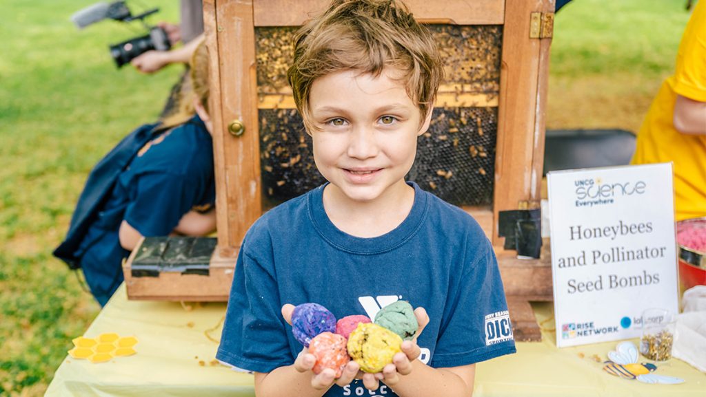 A small boy holds a seed bomb craft he made in front of a beehive.