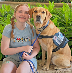 AnnaGrace Berry with braided hair sits on a bench beside a golden retriever wearing a service dog vest. She holds the leash and smiles at the camera while the dog sits calmly, with green plants in the background.