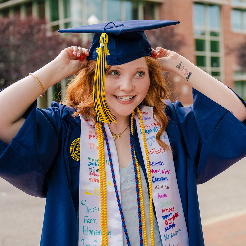 AnnaGrace Berry wearing a navy cap and gown with honor cords adjusts the mortarboard while standing outdoors on campus, with a brick academic building visible behind