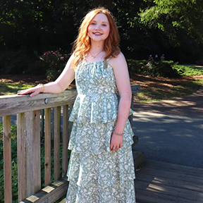 AnnaGrace Berry wearing a sleeveless, tiered green patterned dress stands on a wooden footbridge, resting one hand on the railing, with trees and sunlit greenery in the background.
