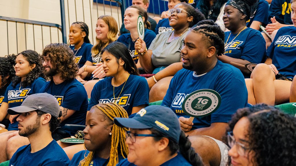 UNCG students sit in bleachers in matching blue shirts.