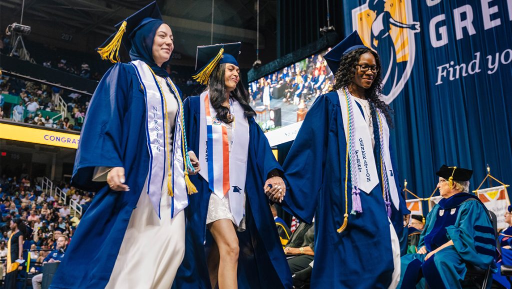 Three UNCG grads walk across the stage holding hands.