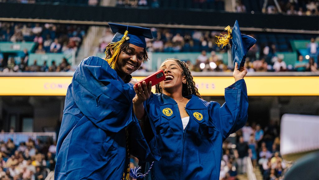 Two UNCG grads smile while looking at a phone.