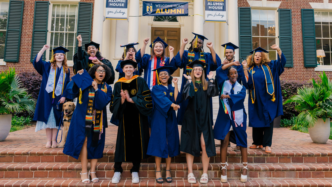 A group of UNCG grads in cap and gowns celebrate on steps outside the Johnson Alumni House.