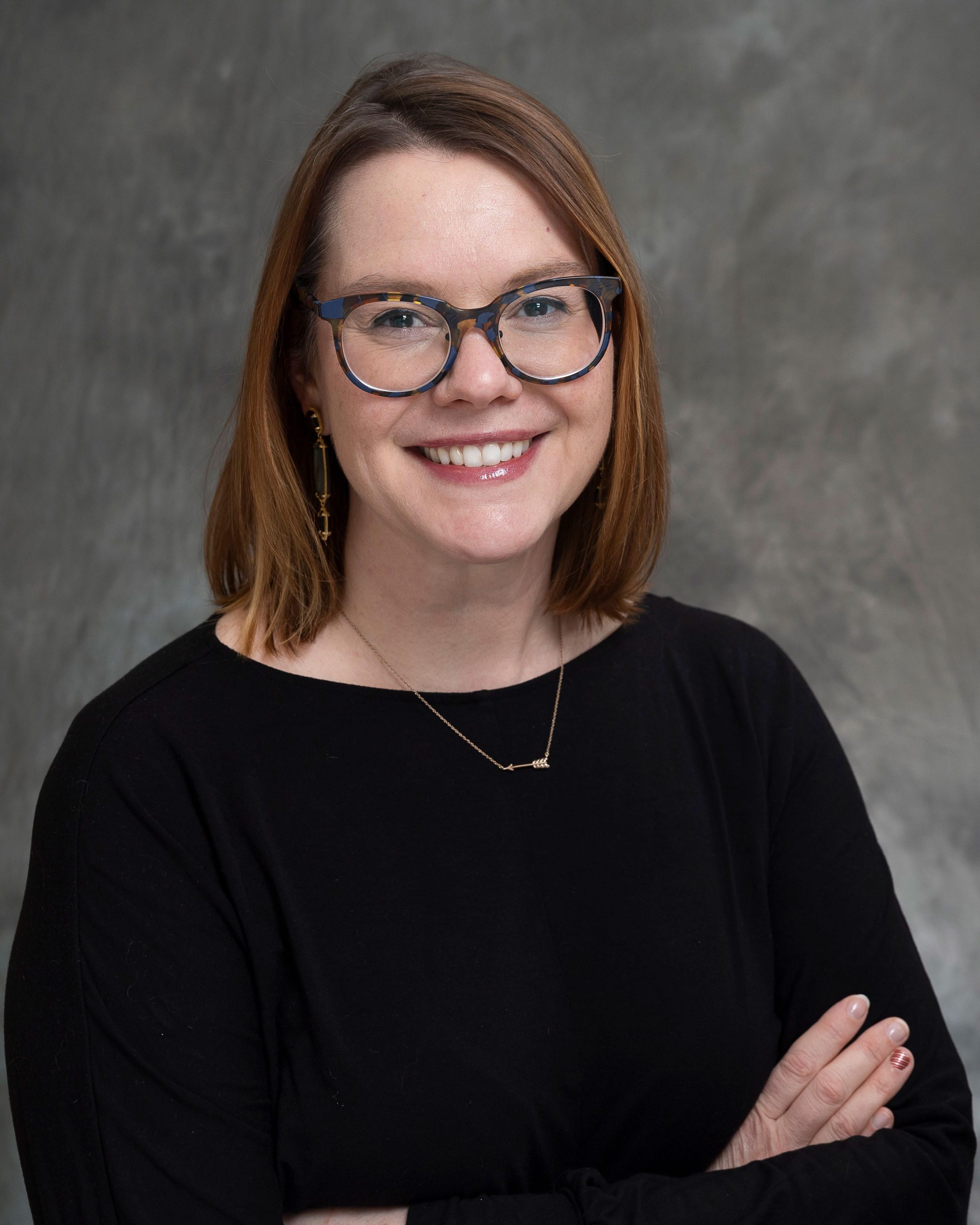 Professional headshot of Adams in a black top with arms crossed, smiling, against a gray background.