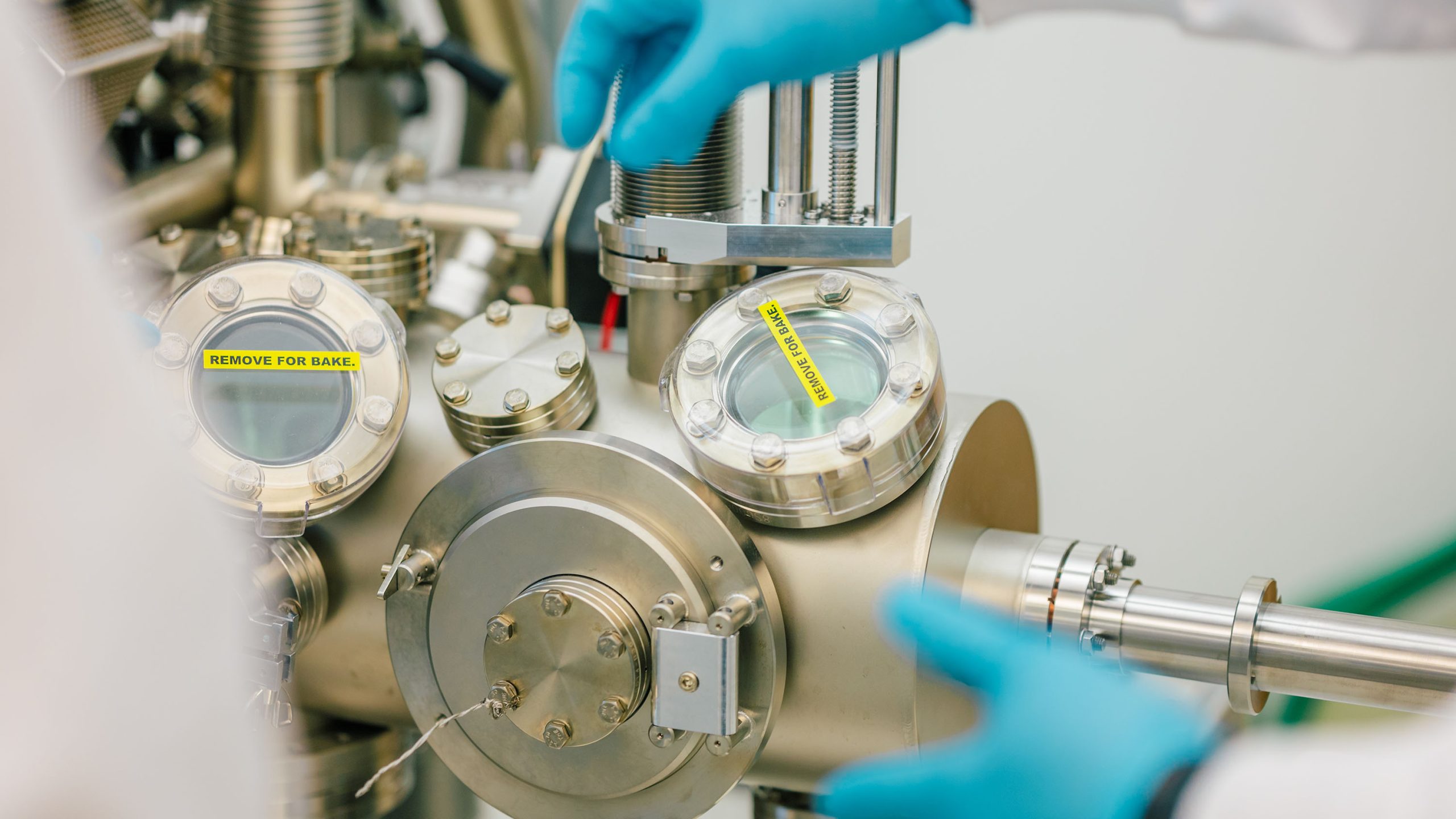 Gloved hands adjust components on a stainless steel vacuum chamber system in a research laboratory.