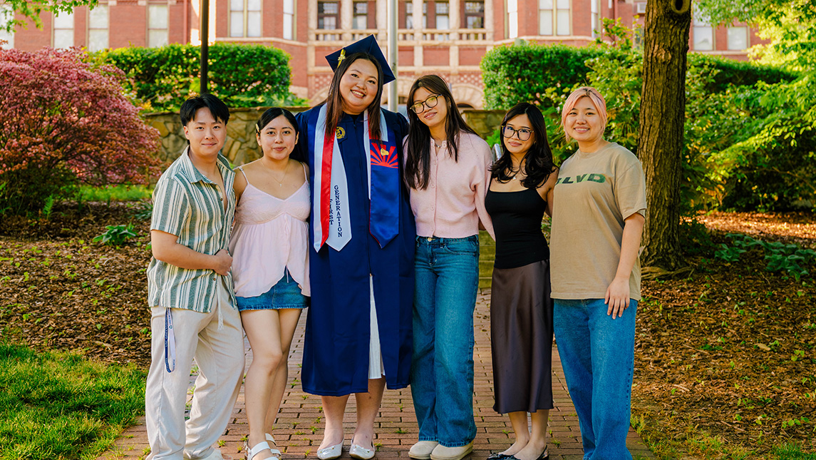 Group of students pose with a graduate in cap and gown in front of Foust.