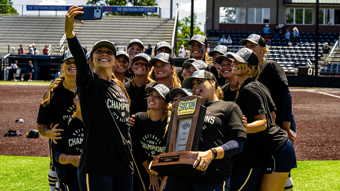 Softball players take a selfie on the field holding a trophy and wearing SoCon championship shirts.