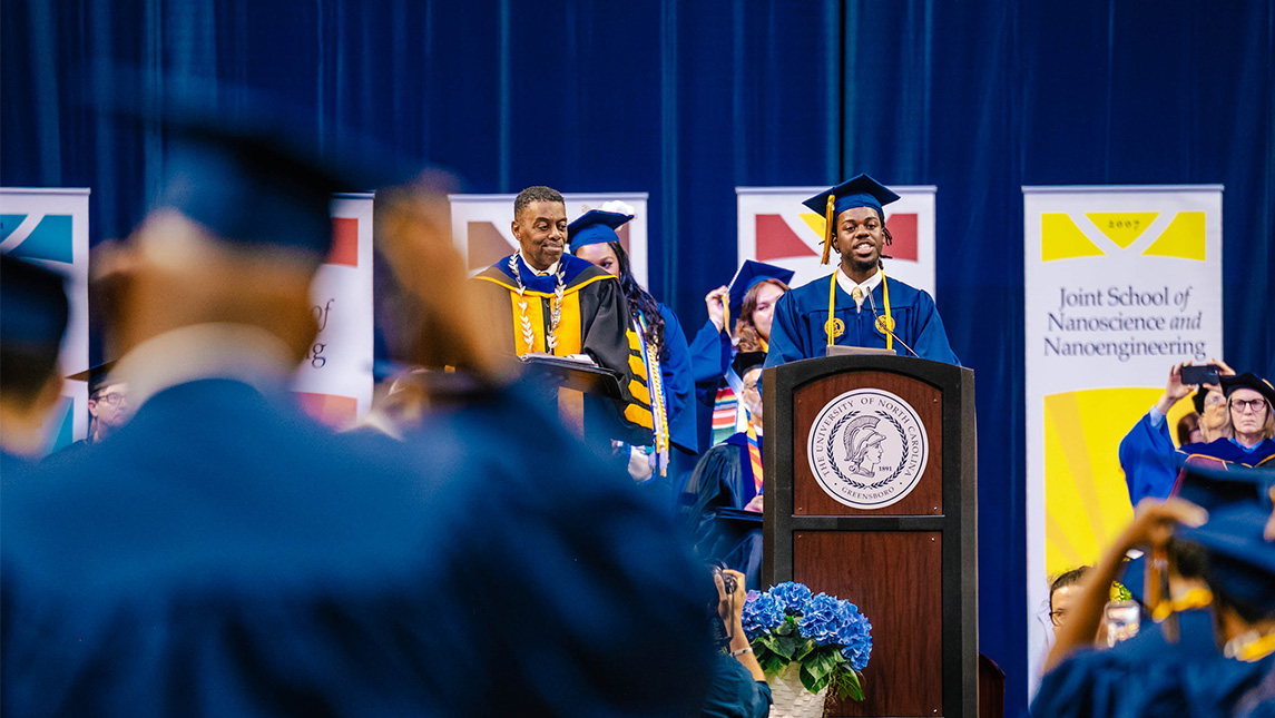 Christian Bartney in his cap and gown speaks at the podium during the UNCG Commencement.