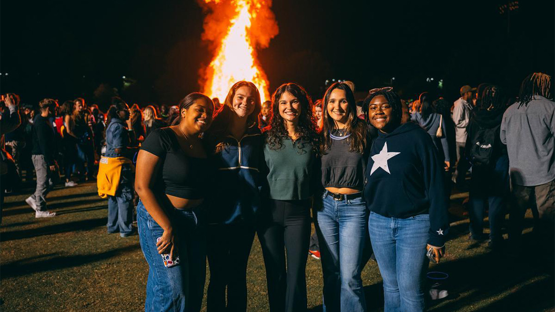 Group of girls smile in front of a bonfire at night.