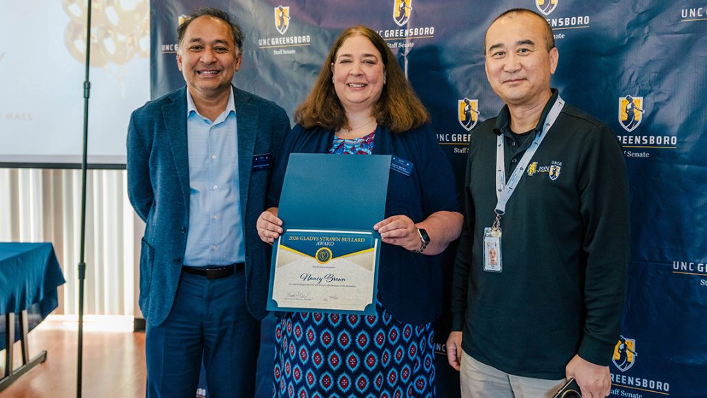 Dean Masud Chowdhury, Nancy Brown, and Yirong Mo pose in front of a UNCG backdrop.