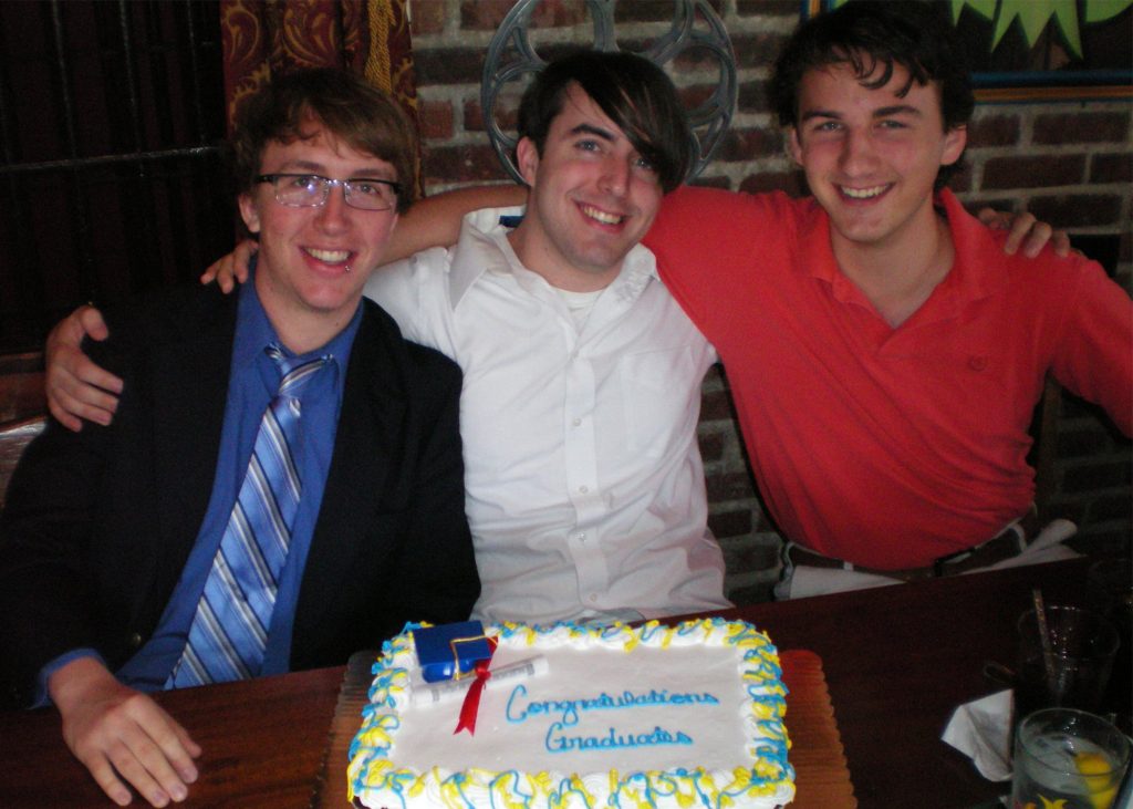 Three men pose in front of a graduation cake.