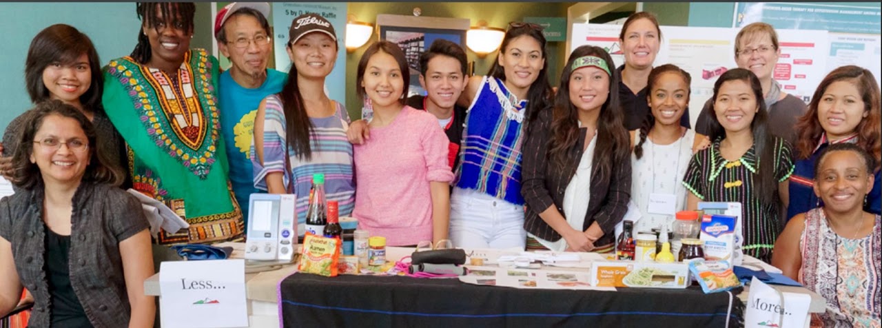 A diverse group of students, faculty, and staff stand together behind a display table during a cultural heritage event. Several participants wear traditional clothing from different cultures, and the table features food items, household products, and informational materials representing cultural traditions. Everyone is smiling and posing for a group photo in an indoor campus setting.