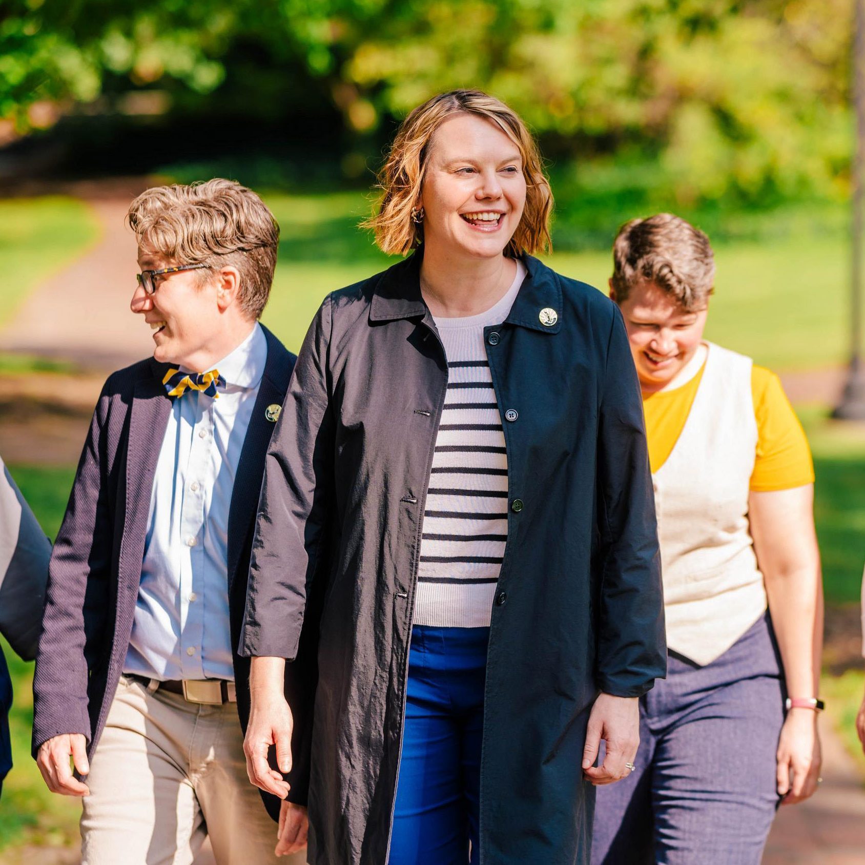 Five people walk together along a sunlit outdoor path surrounded by lush green trees and grass. They are smiling and laughing, conveying a sense of camaraderie.