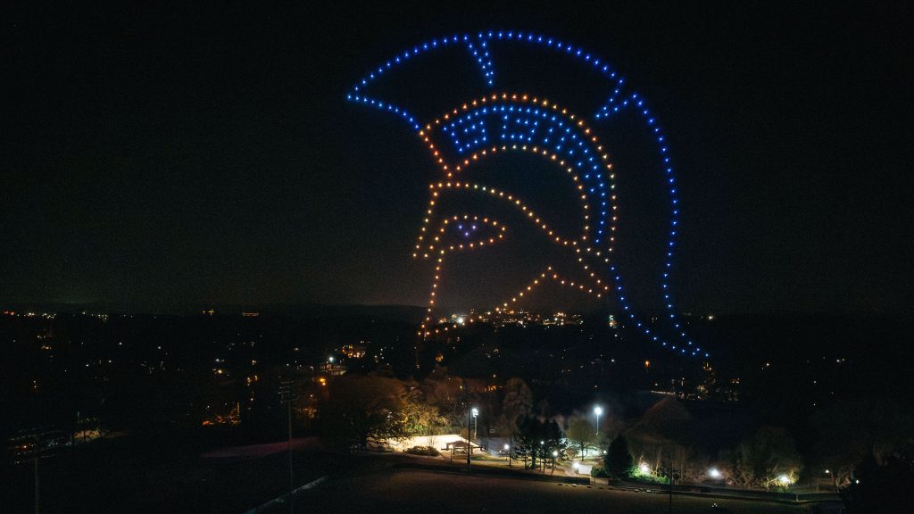 A drone show in the air where the drones have drawn out the Spartan helmet.