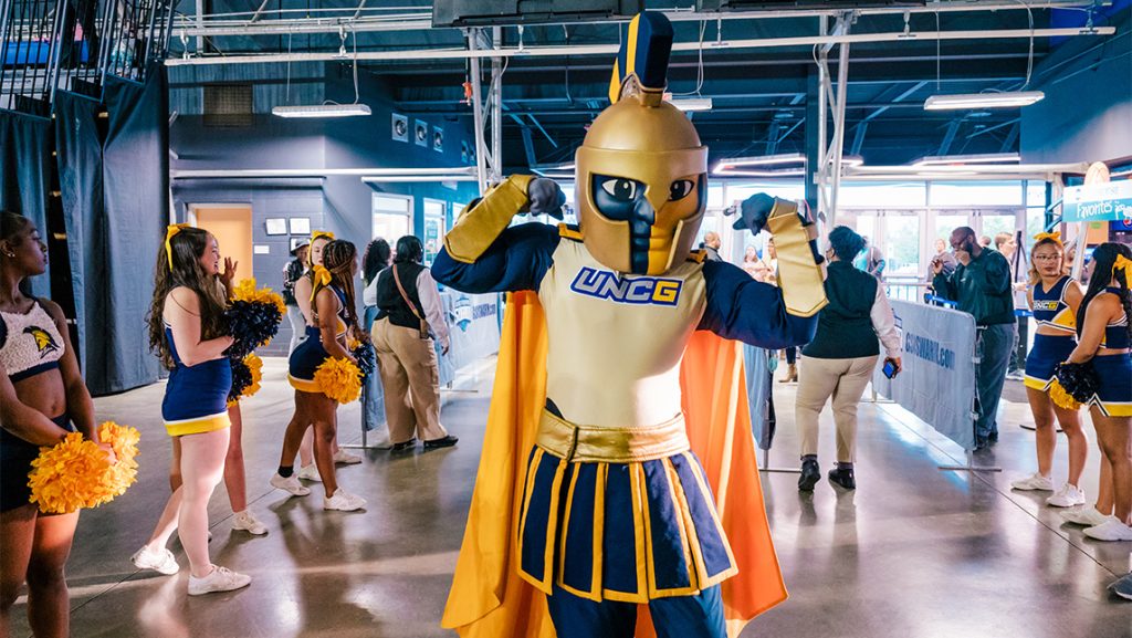 UNCG mascot Spiro with cheerleaders inside a basketball stadium.